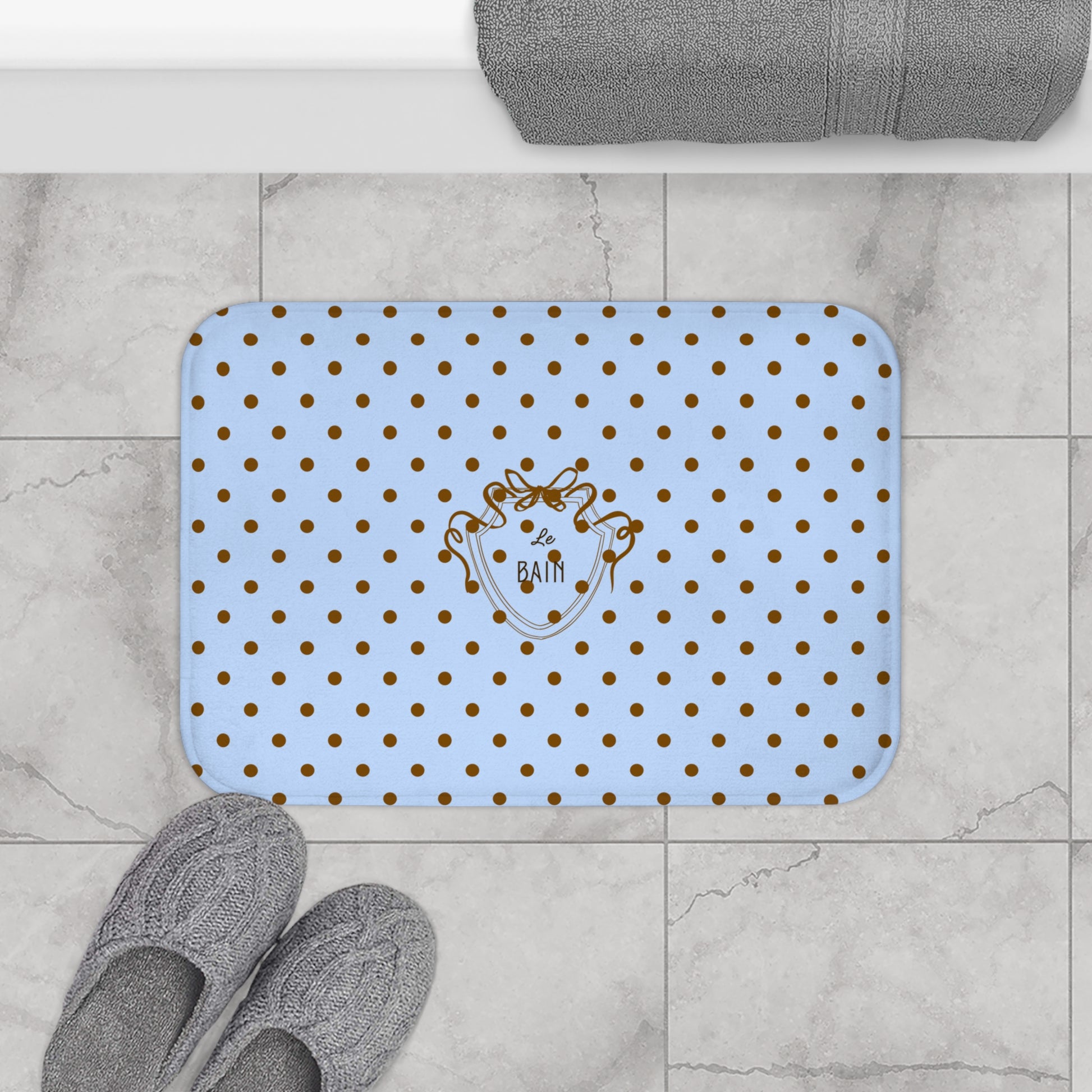 Blue bath mat with gold polka dots and a logo on a tiled bathroom floor.
