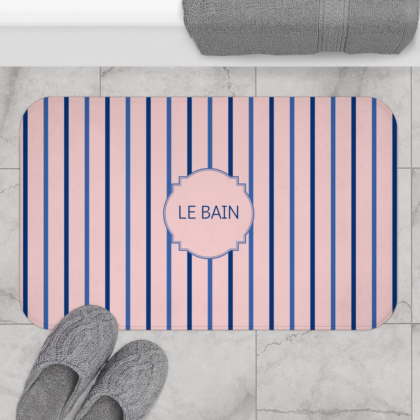 A rectangular bath mat with rounded corners, featuring vertical pink and navy blue stripes and a central decorative label that reads 'LE BAIN'. The mat is styled in a bathroom setting on a light gray marble tile floor, with gray slippers at the bottom left and a folded gray towel at the top.