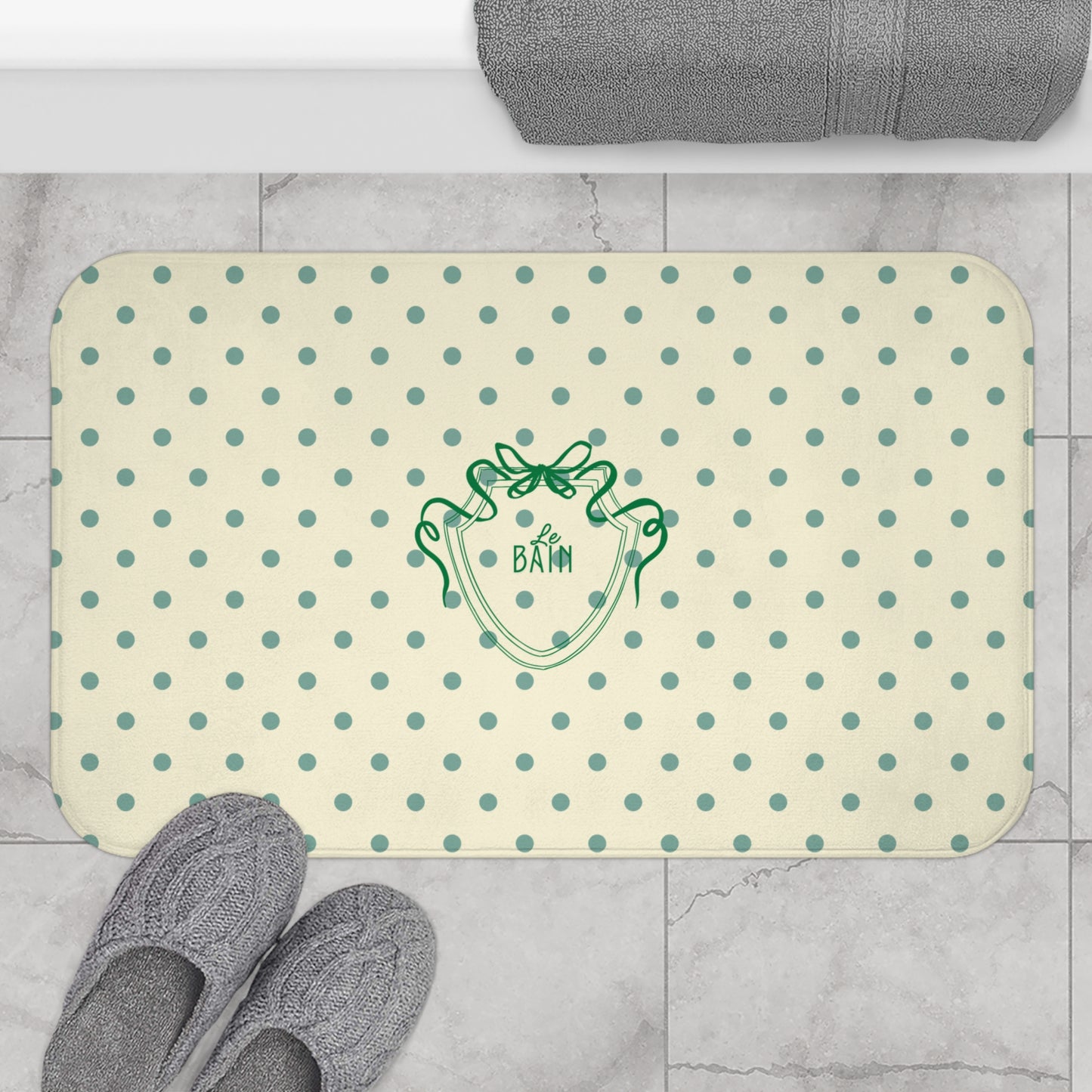Beige bath mat with green polka dots and logo on a tiled bathroom floor.