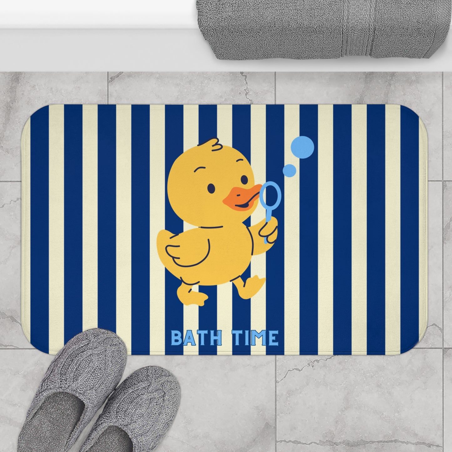A rectangular bath mat with rounded corners, featuring a yellow cartoon duck blowing blue bubbles on a navy and cream vertical stripe background. The words 'BATH TIME' are printed in blue near the bottom.