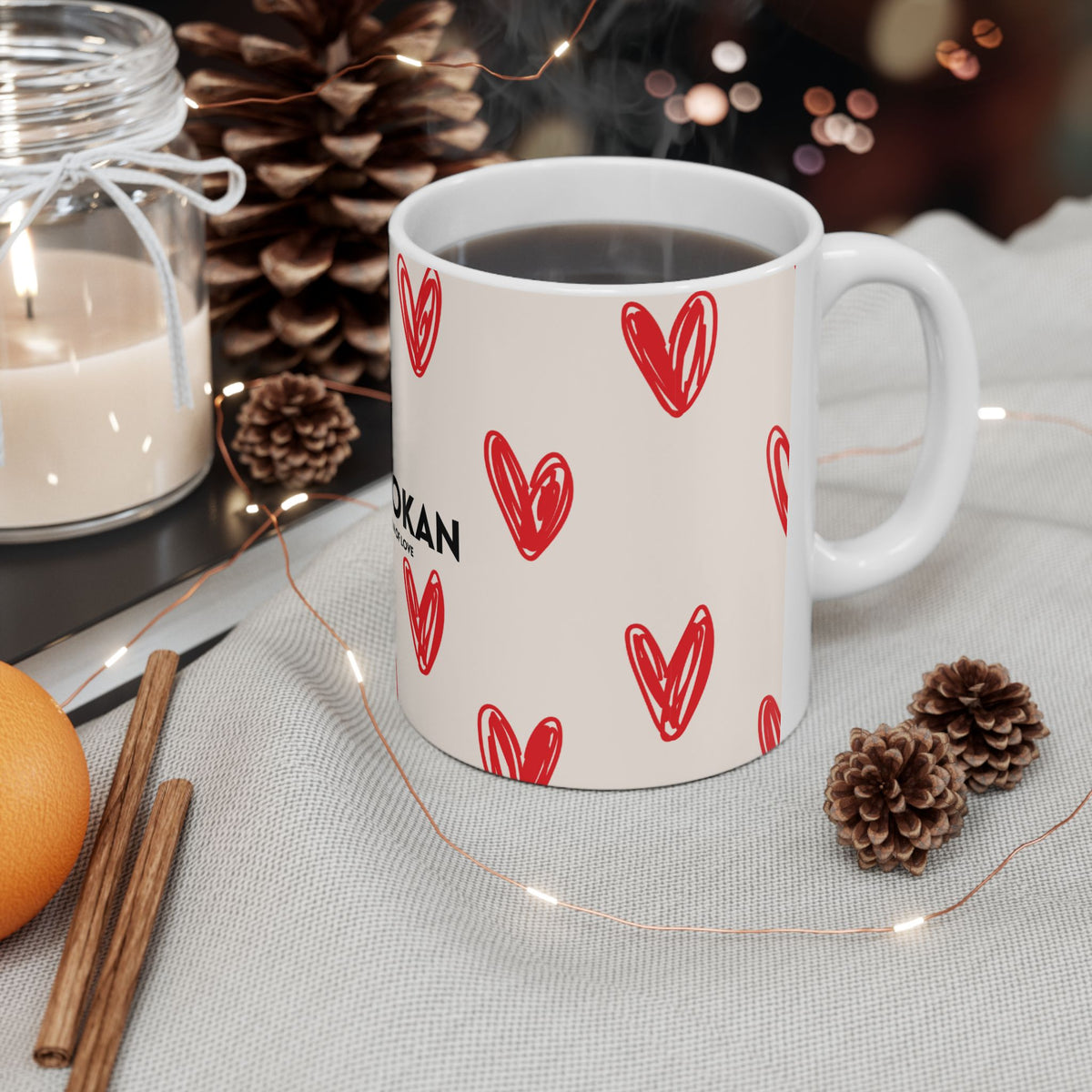 A white ceramic mug filled with a dark beverage, decorated with a pattern of red hand-drawn hearts and partial black text, styled on a light fabric surface with pinecones, fairy lights, a candle, cinnamon sticks, and an orange.