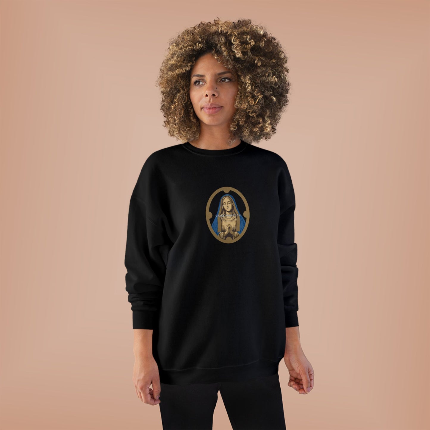 A woman with a large curly afro hairstyle wearing a black crewneck sweatshirt with a circular graphic of the Virgin Mary on the chest, set against a solid peach-colored background.
