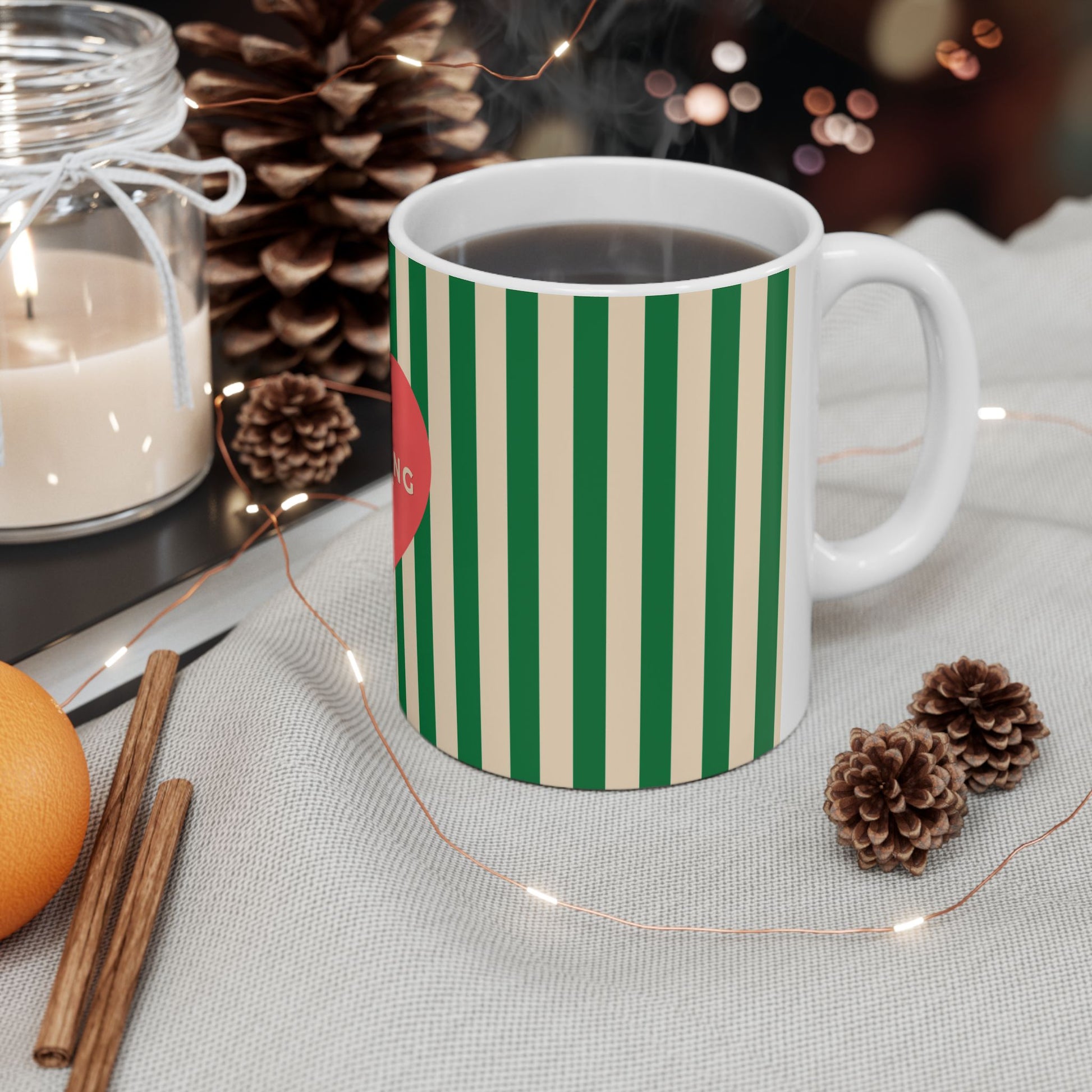 A white ceramic mug with vertical green and cream stripes, filled with a dark beverage, styled on a light fabric surface with pinecones, fairy lights, cinnamon sticks, an orange, and a candle in the background.