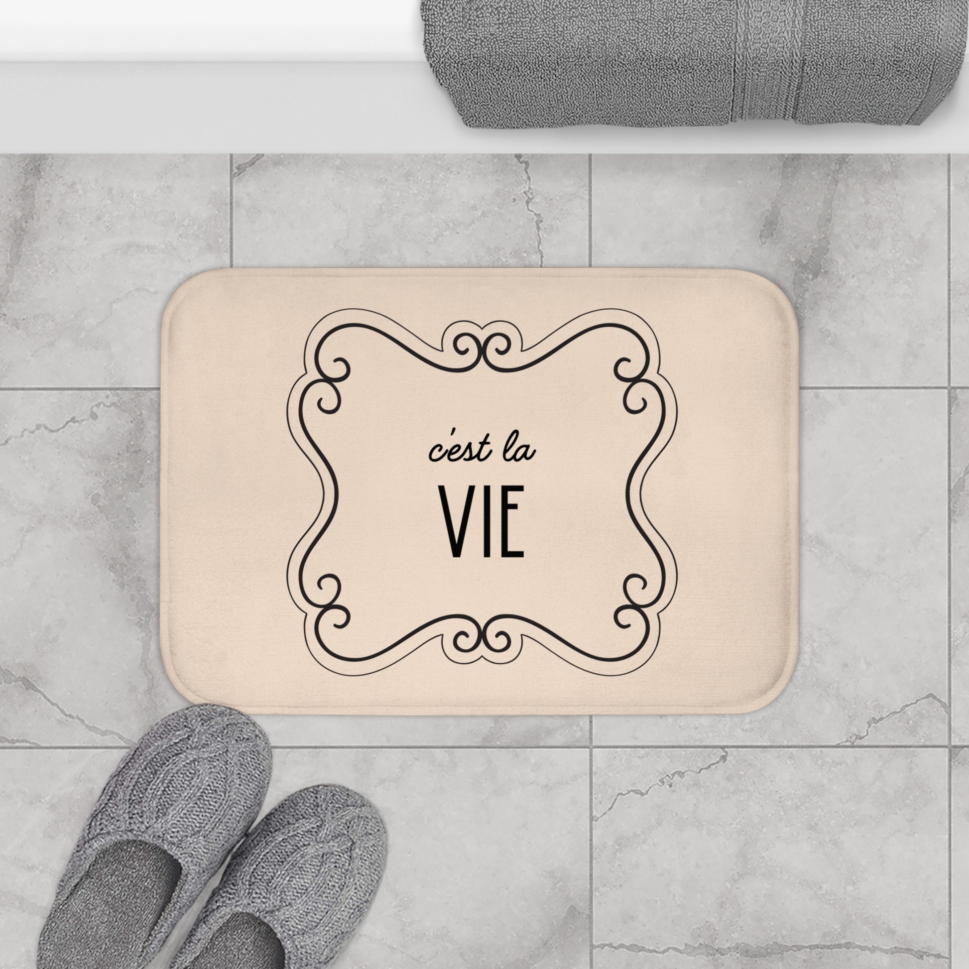Beige doormat with 'c'est la VIE' text on a marble floor with slippers and a towel.