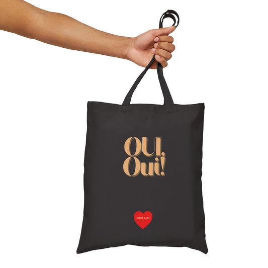 A black canvas tote bag with gold 'Oui, Oui!' text and a small red heart graphic, held by a person's arm against a plain white background.