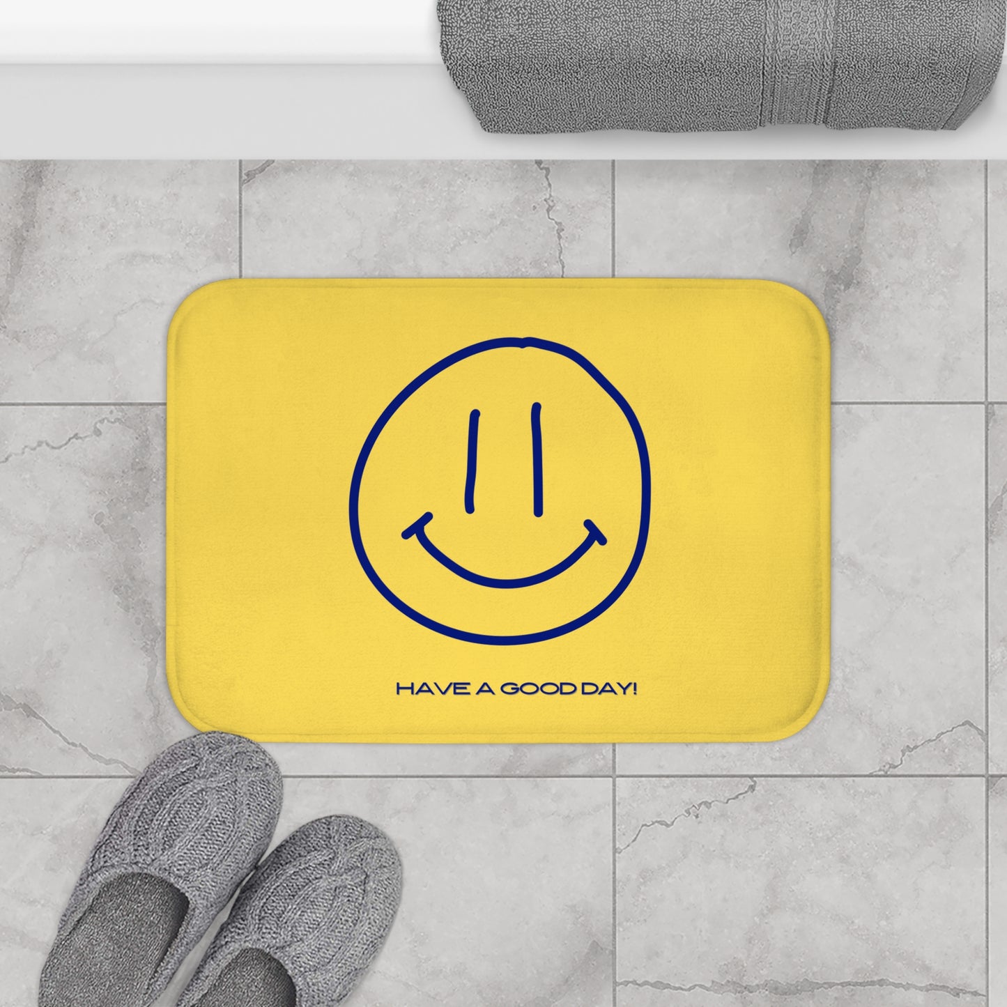 Yellow bath mat with blue smiley face and text 'Have a good day!' on a marble floor.