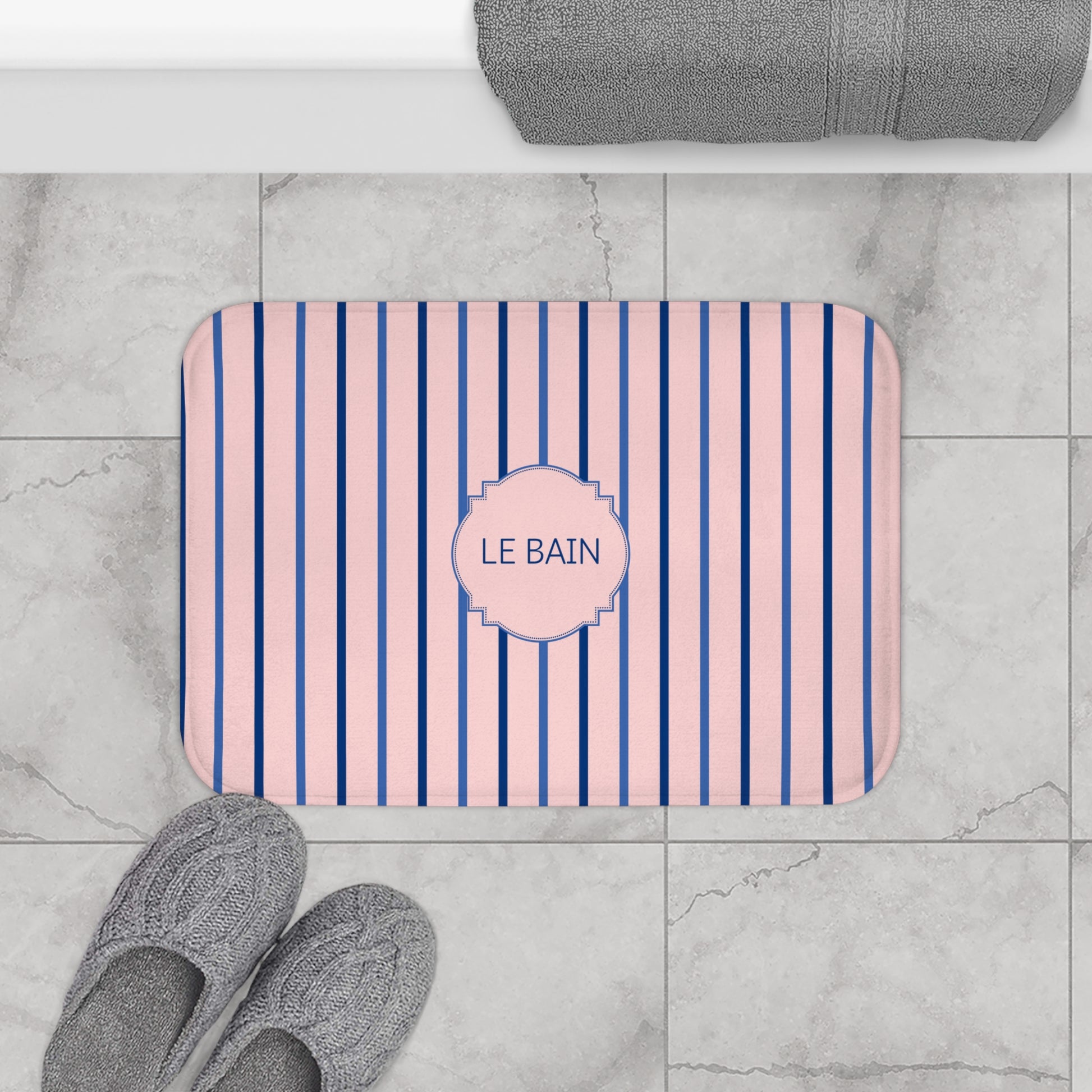 A rectangular bath mat with pink and navy blue vertical stripes and a central pink label reading 'LE BAIN', shown on a light gray marble tile floor with gray towels and slippers nearby.