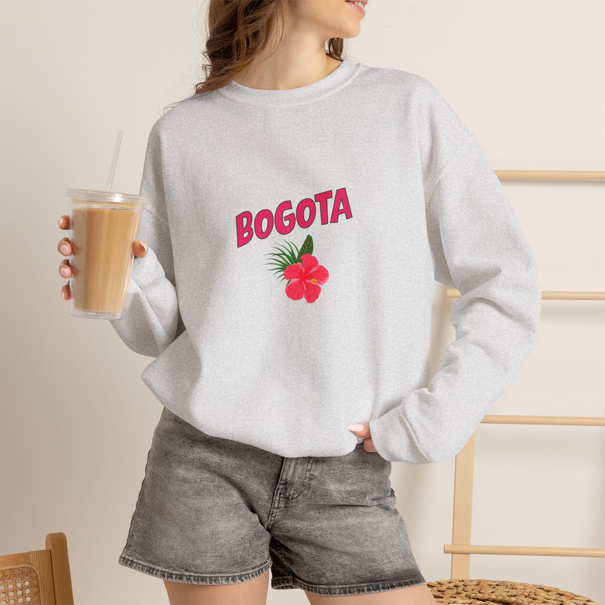 Person wearing a gray sweatshirt with 'BOGOTA' and a flower design, holding a drink.