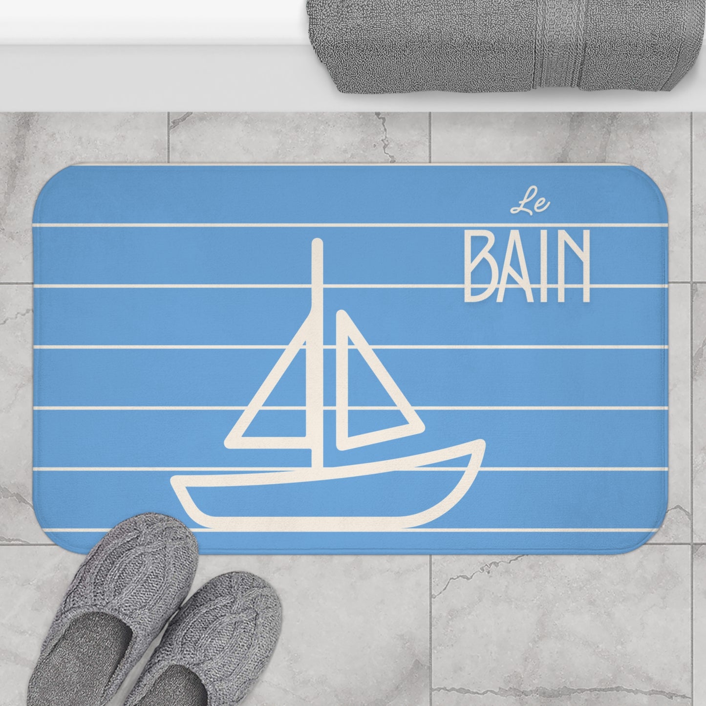 Blue bath mat with white sailboat design and 'Le Bain' text on a marble floor.