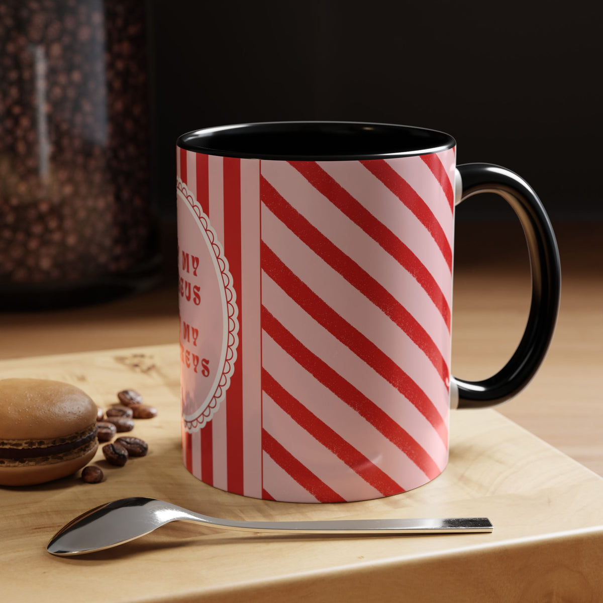 A ceramic coffee mug with a red and white diagonal stripe pattern and a black handle, sitting on a wooden surface next to a spoon with coffee beans and a macaron in the background.