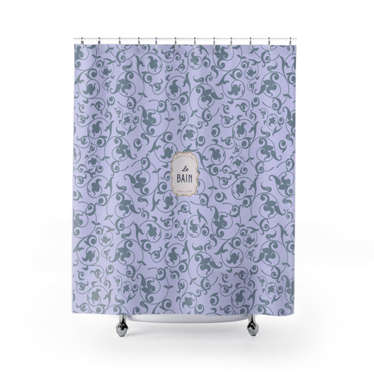 A lavender purple shower curtain with a repeating gray floral scroll pattern and a central cream-colored decorative label that reads 'BATH'. The curtain is shown hanging from silver hooks over a white tub rim with small silver feet visible at the bottom, set against a plain white background.