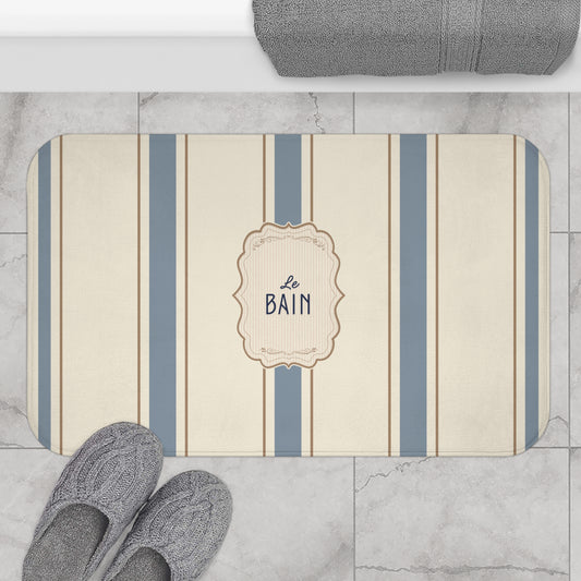 Striped bath mat with 'Le Bain' text on a marble floor with slippers and towel.