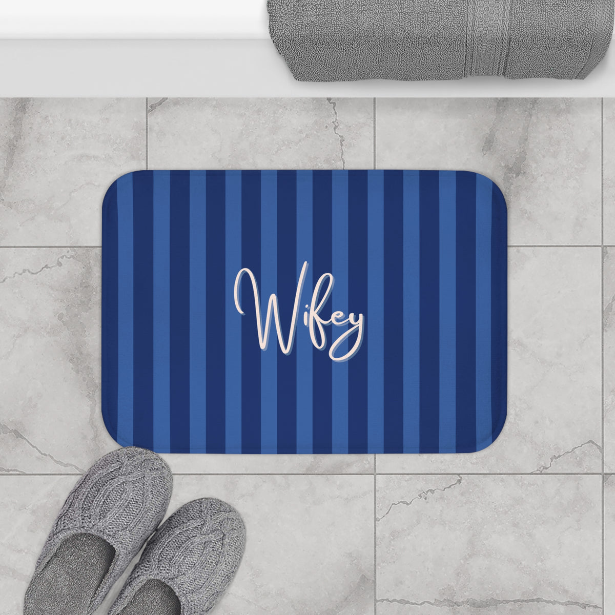 A rectangular bath mat with rounded corners, featuring vertical navy and medium blue stripes and the name 'Wifey' in white script at the center. The mat is shown on a light gray marble tile floor with gray slippers and a folded gray towel nearby.