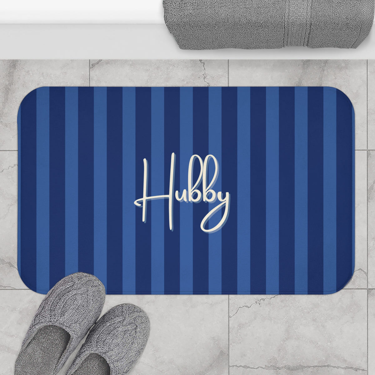 A rectangular bath mat with rounded corners, featuring vertical navy and medium blue stripes and the name 'Hubby' in white script. The mat is shown on a light marble tile floor with gray slippers and a folded gray towel nearby.