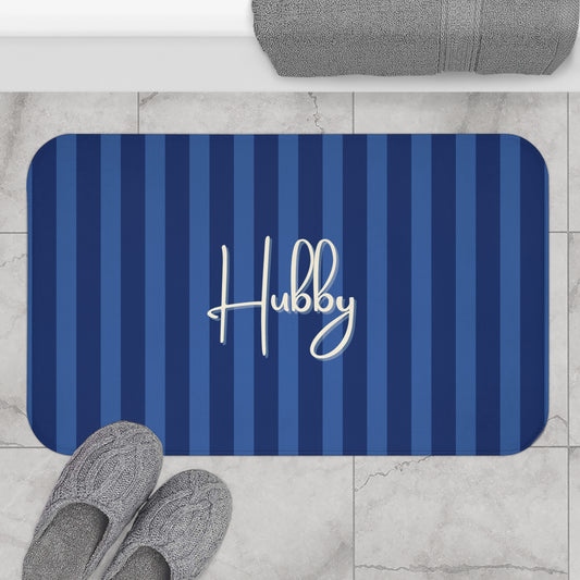 A rectangular bath mat with rounded corners, featuring vertical navy and medium blue stripes and the name 'Hubby' in white script. The mat is shown on a light marble tile floor with gray slippers and a folded gray towel nearby.