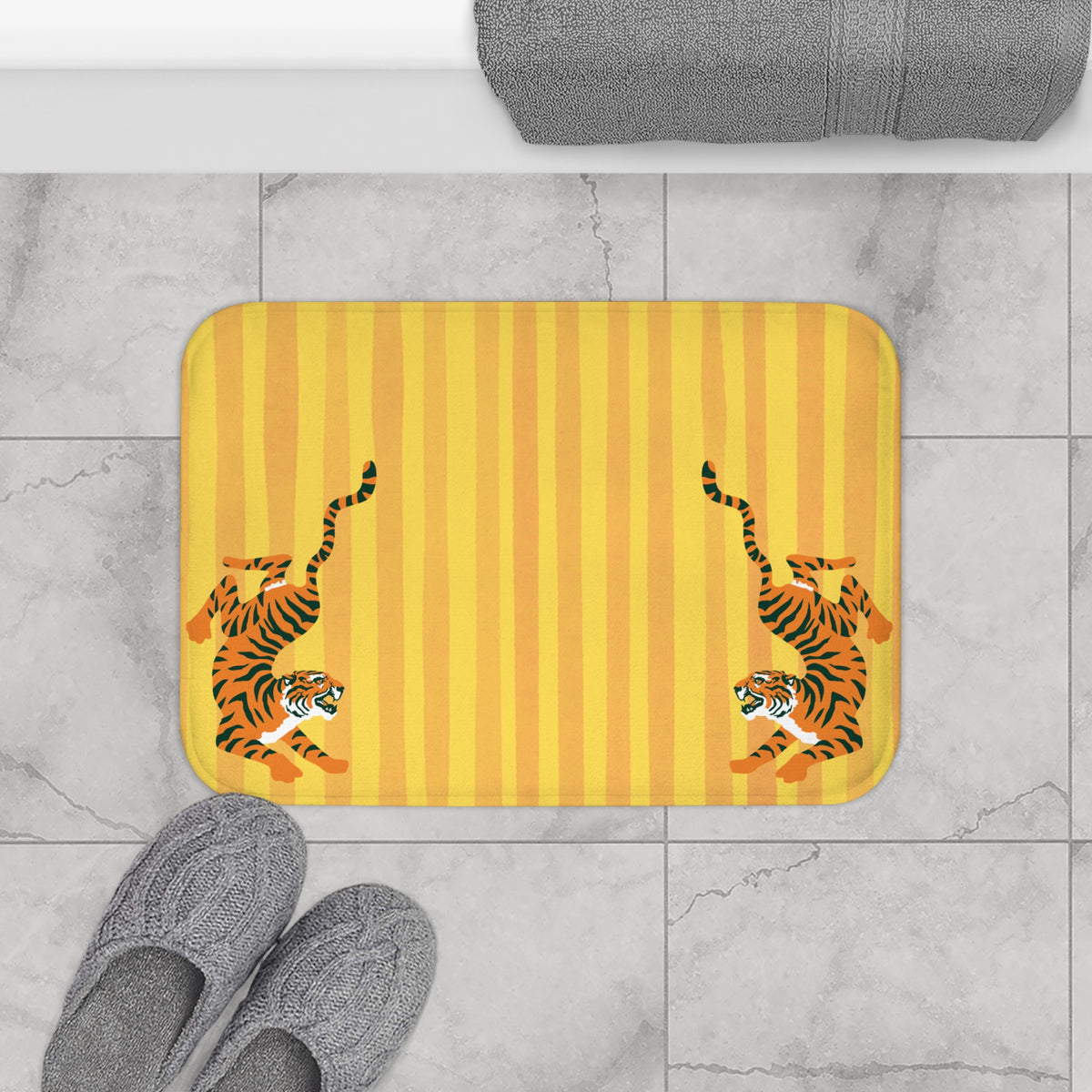 A rectangular bath mat with a yellow and orange striped background and two cartoon tigers lying down, shown on a light gray tiled floor with gray towels and slippers nearby.