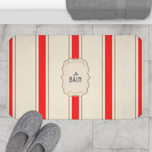 Bath mat with red stripes and 'Le Bain' text on a marble floor with slippers and a towel.
