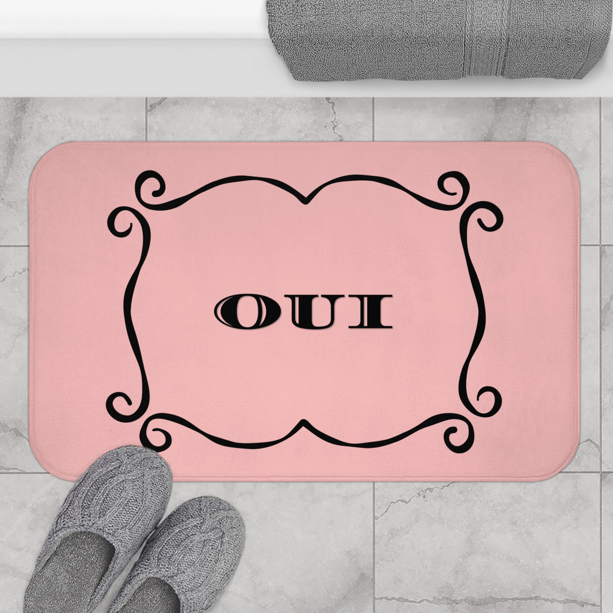 A pink rectangular bath mat with rounded corners, featuring a black ornate border and the word 'OUI' in the center. The mat is shown on a light gray marble tile floor with gray slippers and a folded gray towel nearby.
