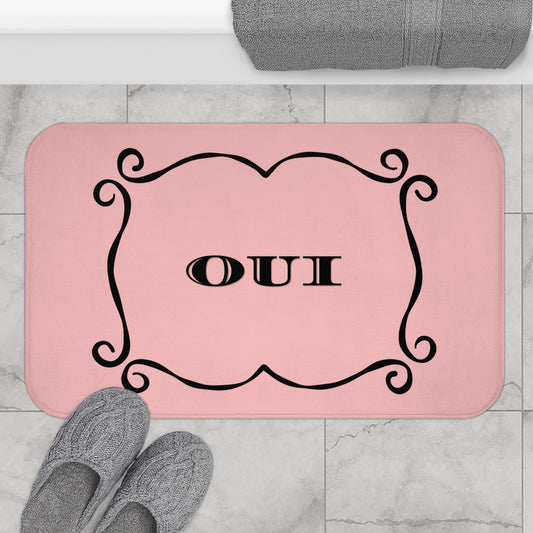 A pink rectangular bath mat with rounded corners, featuring a black ornate border and the word 'OUI' in the center. The mat is shown on a light gray marble tile floor with gray slippers and a folded gray towel nearby.