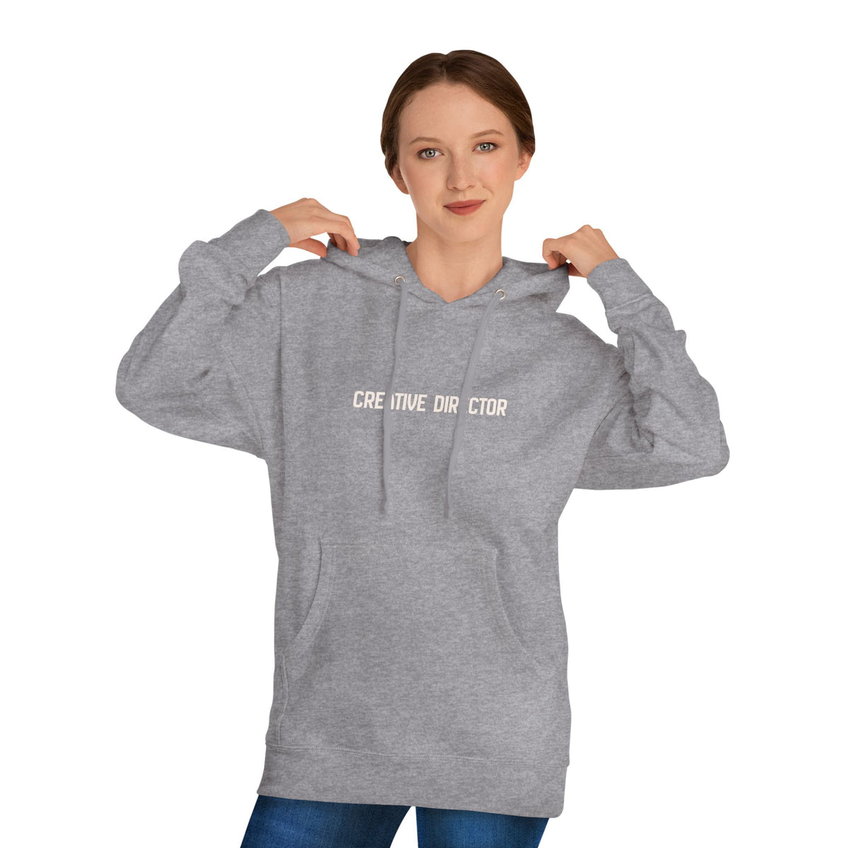 A woman wearing a heather gray pullover hoodie with the words 'CREATIVE DIRECTOR' printed in white across the chest, posed against a solid white background.