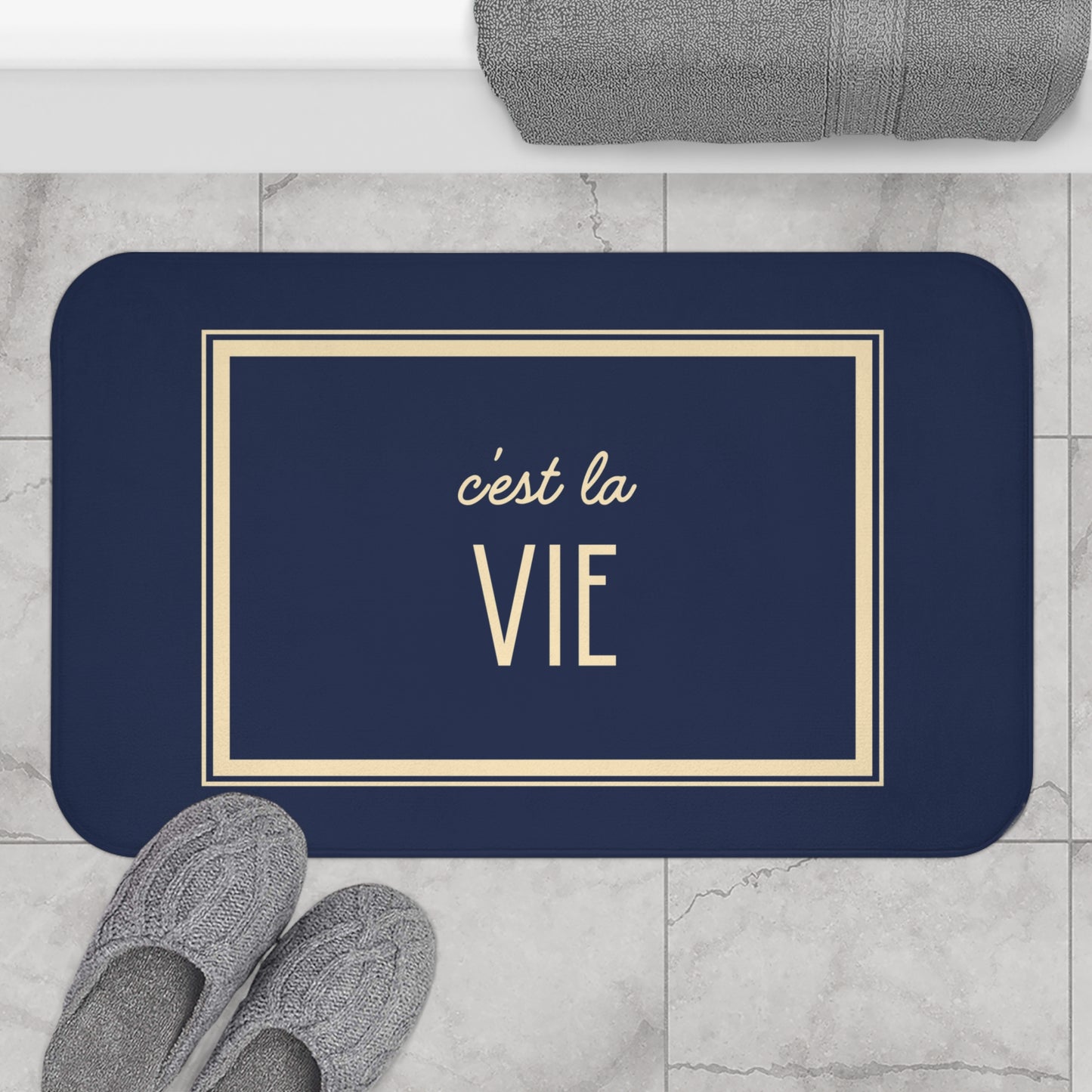 Navy blue mat with 'c'est la Vie' text on a marble floor with slippers and towel.