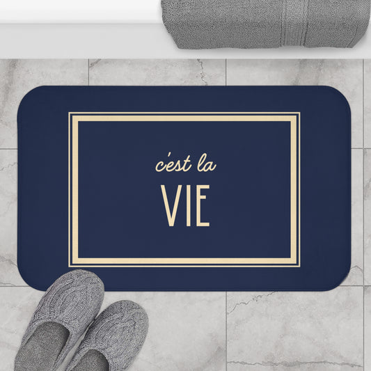Navy blue mat with 'c'est la Vie' text on a marble floor with slippers and towel.