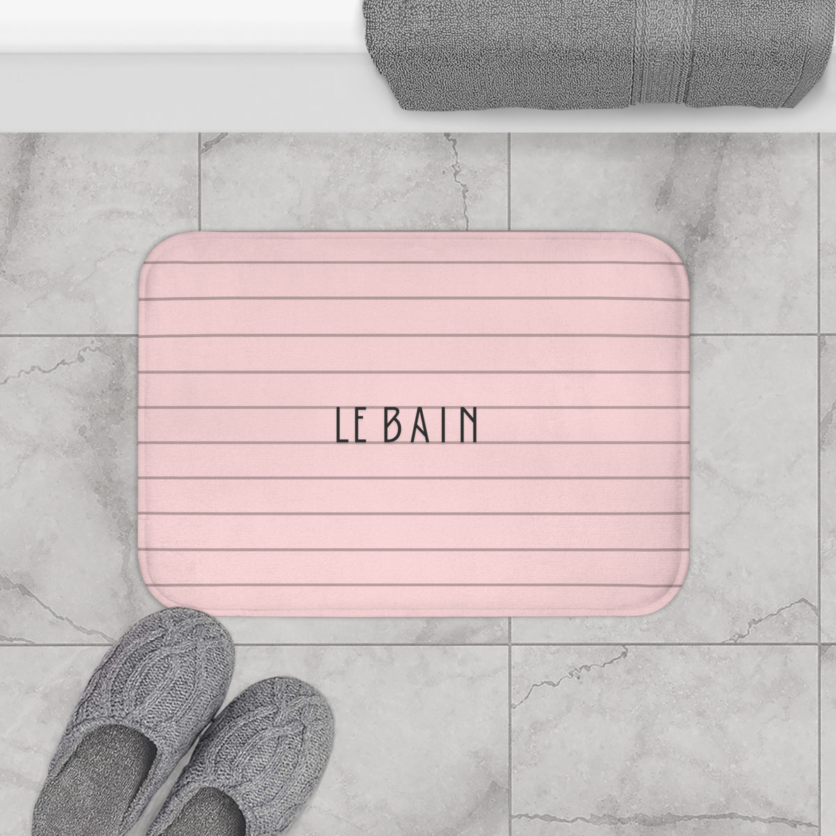 A pink bath mat with horizontal stripes and the text 'LE BAIN' in the center, shown on a gray marble tile floor with gray slippers and a folded towel nearby.