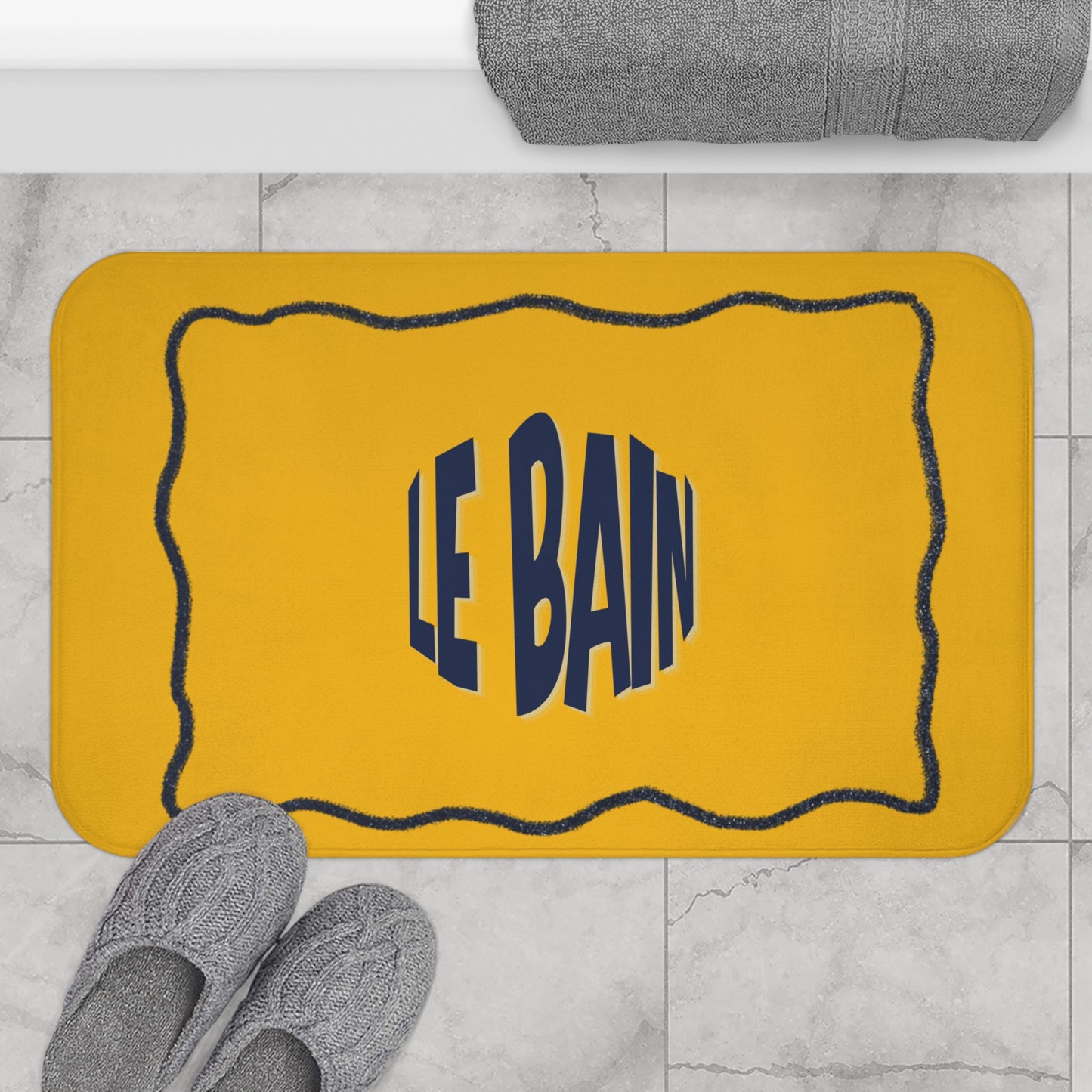 Yellow bath mat with 'LE BAIN' text on a marble floor with gray slippers and towel.