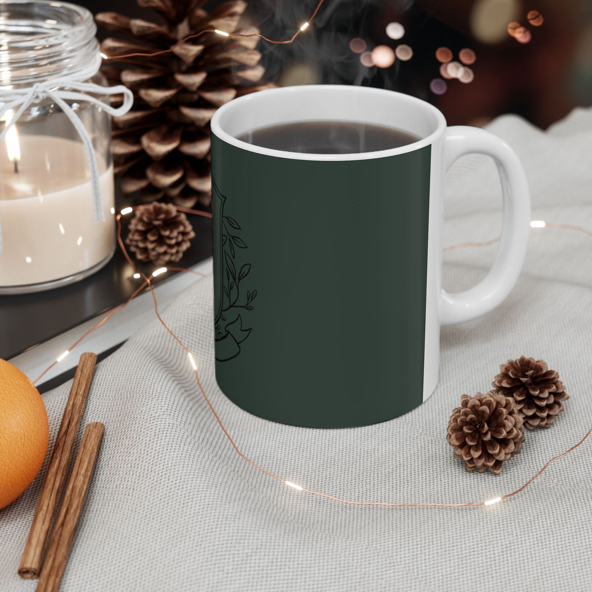 A white ceramic mug filled with a dark beverage, featuring a dark green front panel with a black botanical line drawing. The mug is styled on a light fabric surface with warm holiday props including pinecones, fairy lights, cinnamon sticks, an orange, and a lit candle in a glass jar.