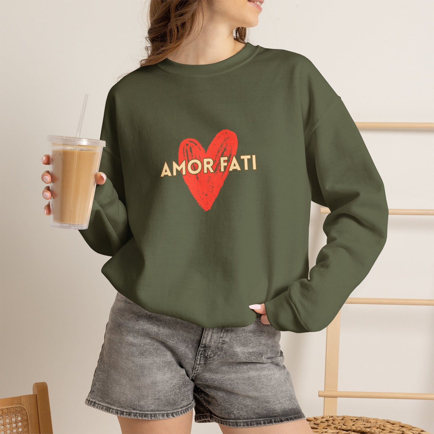 A woman wearing an olive green crewneck sweatshirt with a red heart and 'AMOR FATI' text graphic, paired with gray denim shorts and holding an iced coffee.