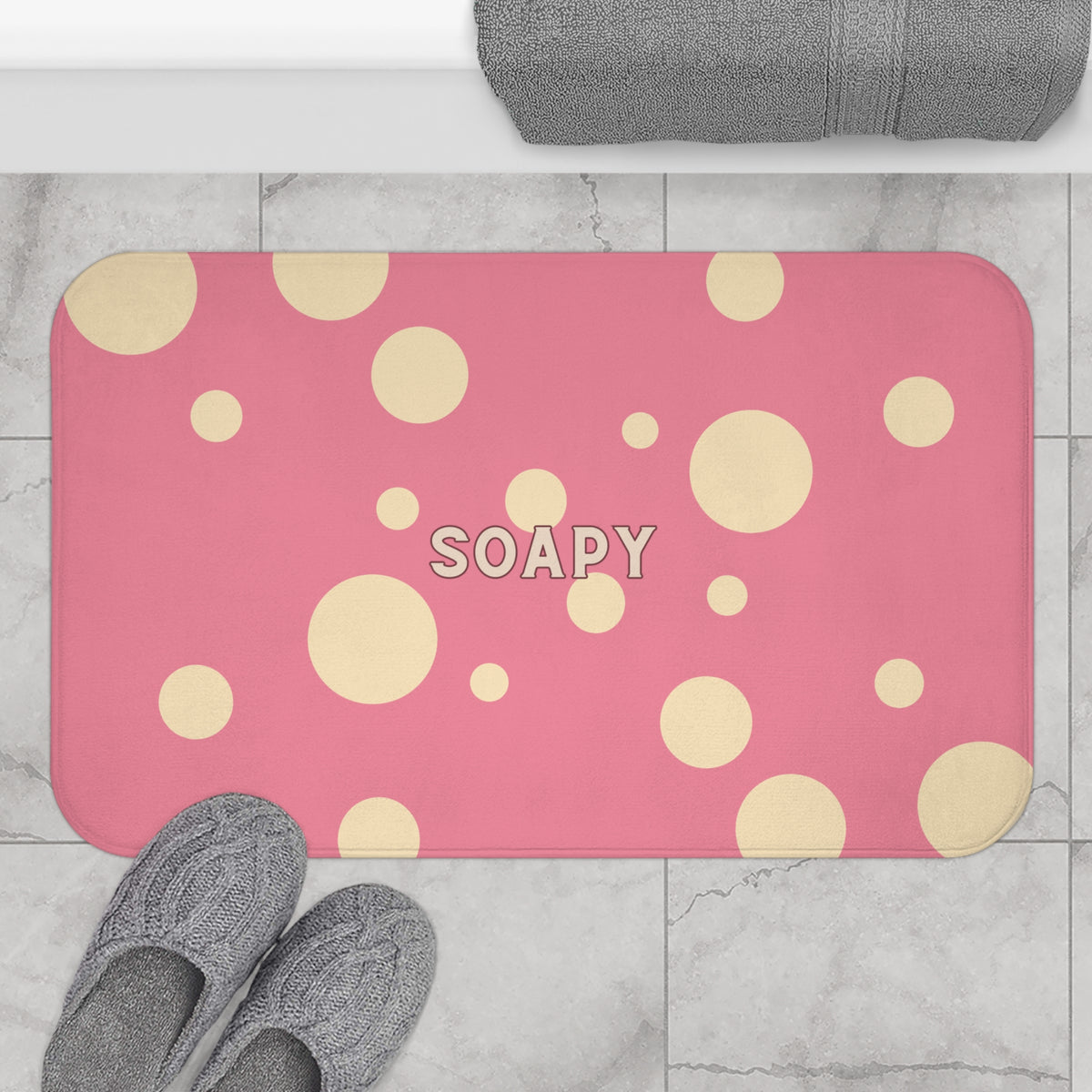 A pink rectangular bath mat with rounded corners, decorated with scattered beige polka dots and the word 'SOAPY' in the center, shown on a light marble floor with gray towels and slippers nearby.