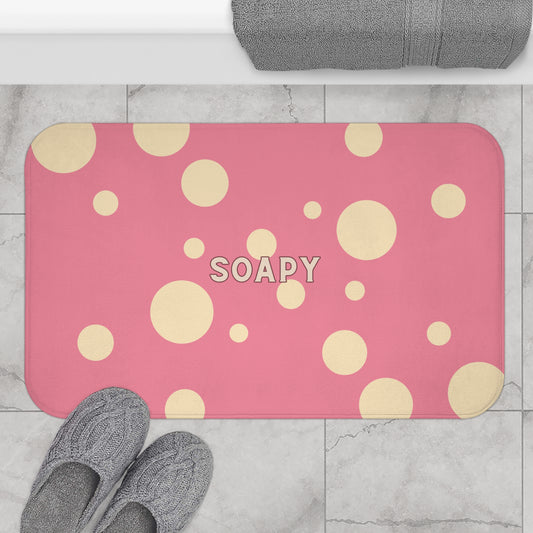 A pink rectangular bath mat with rounded corners, decorated with scattered beige polka dots and the word 'SOAPY' in the center, shown on a light marble floor with gray towels and slippers nearby.