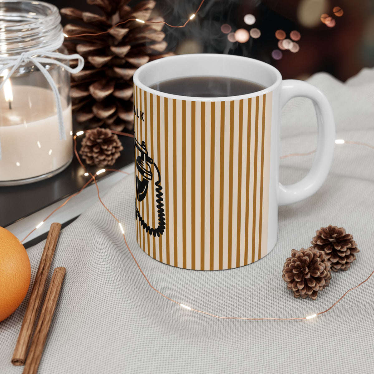 A white ceramic mug with vertical mustard-yellow stripes and a black graphic print, filled with a dark beverage and set on a light fabric surface with pinecones, a candle jar, cinnamon sticks, and warm string lights.
