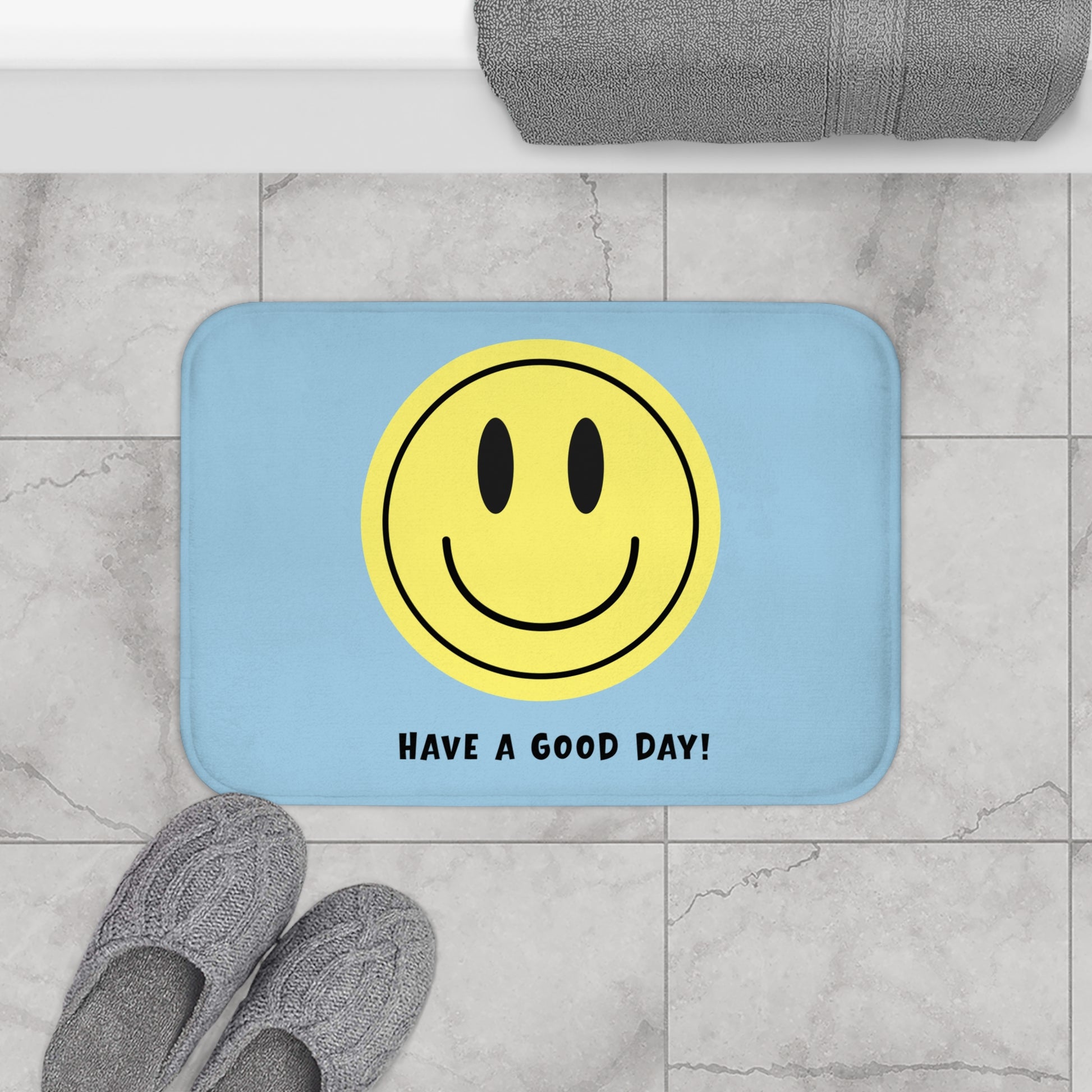 Light blue bath mat with yellow smiley face and 'Have a Good Day!' text on a marble floor.