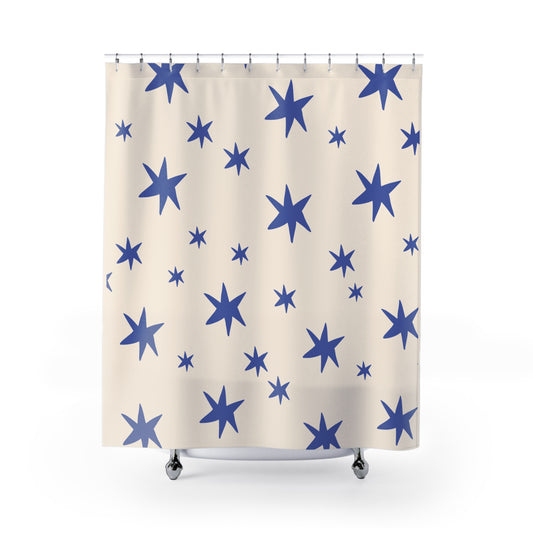 A shower curtain with a pattern of blue stars of various sizes on a warm cream background, hanging from silver rings on a white background.