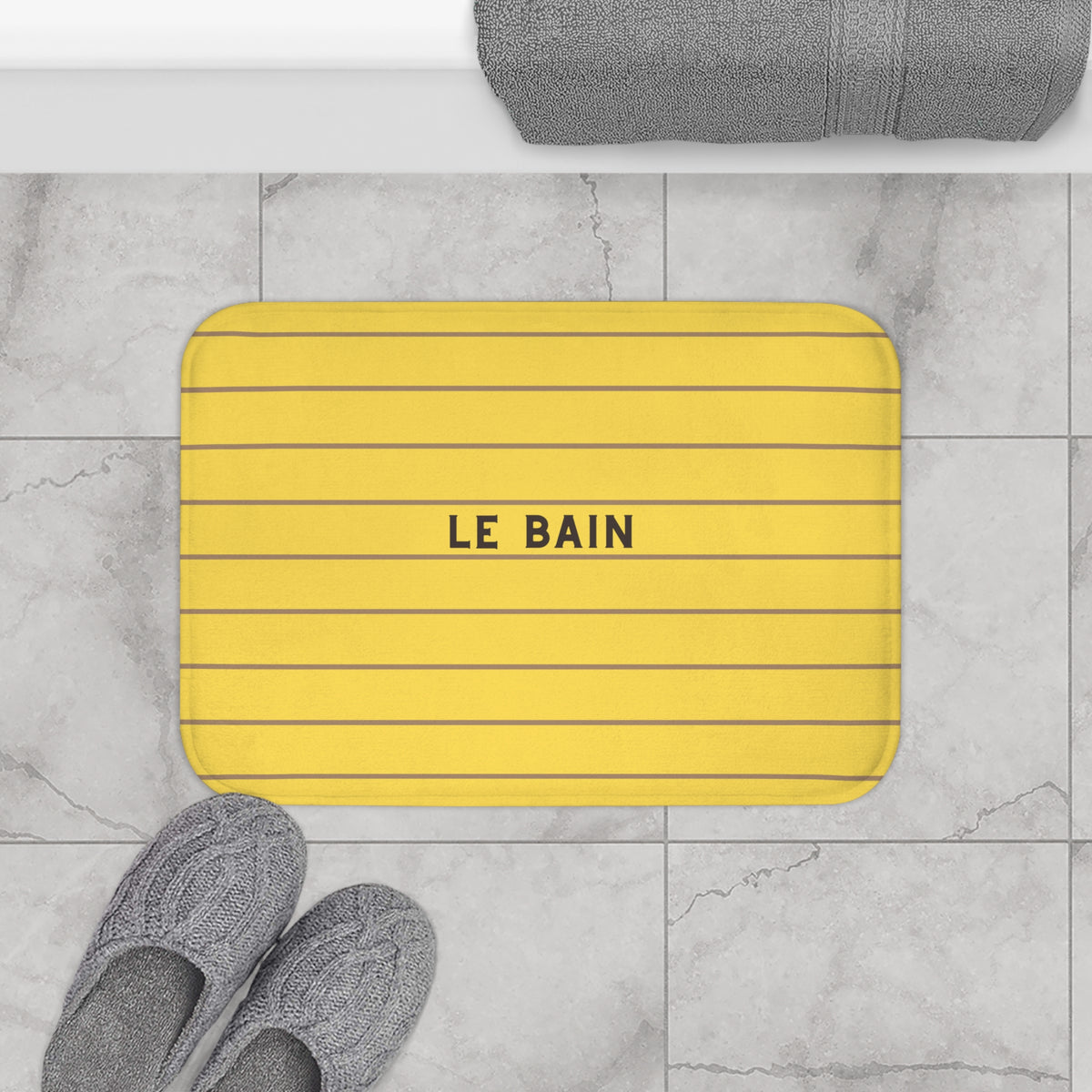 A yellow rectangular bath mat with rounded corners, thin dark horizontal lines, and the words 'LE BAIN' in black at the center, placed on a light gray marble tile floor. Gray towels and slippers are partially visible around it.