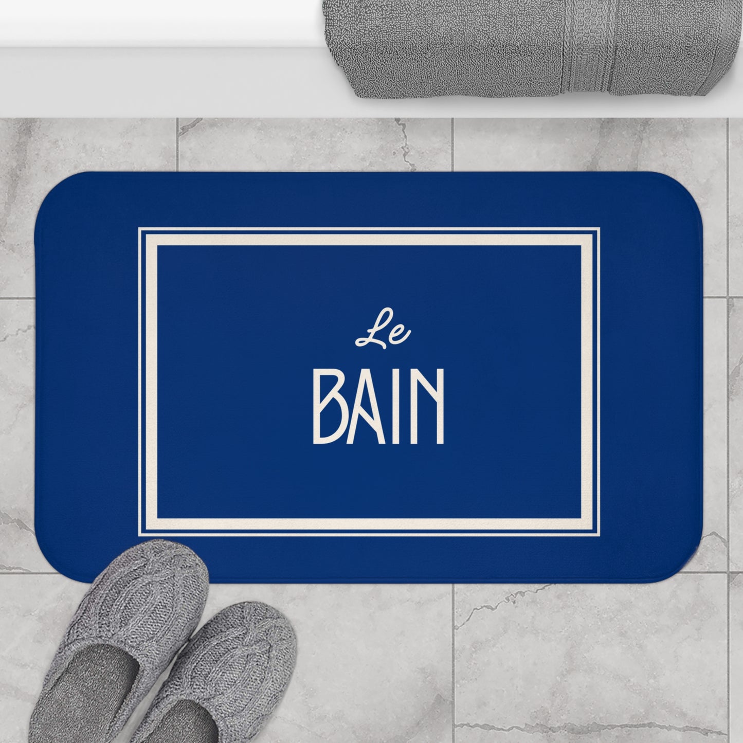 Blue bath mat with 'Le Bain' text on a marble floor with slippers and towel.