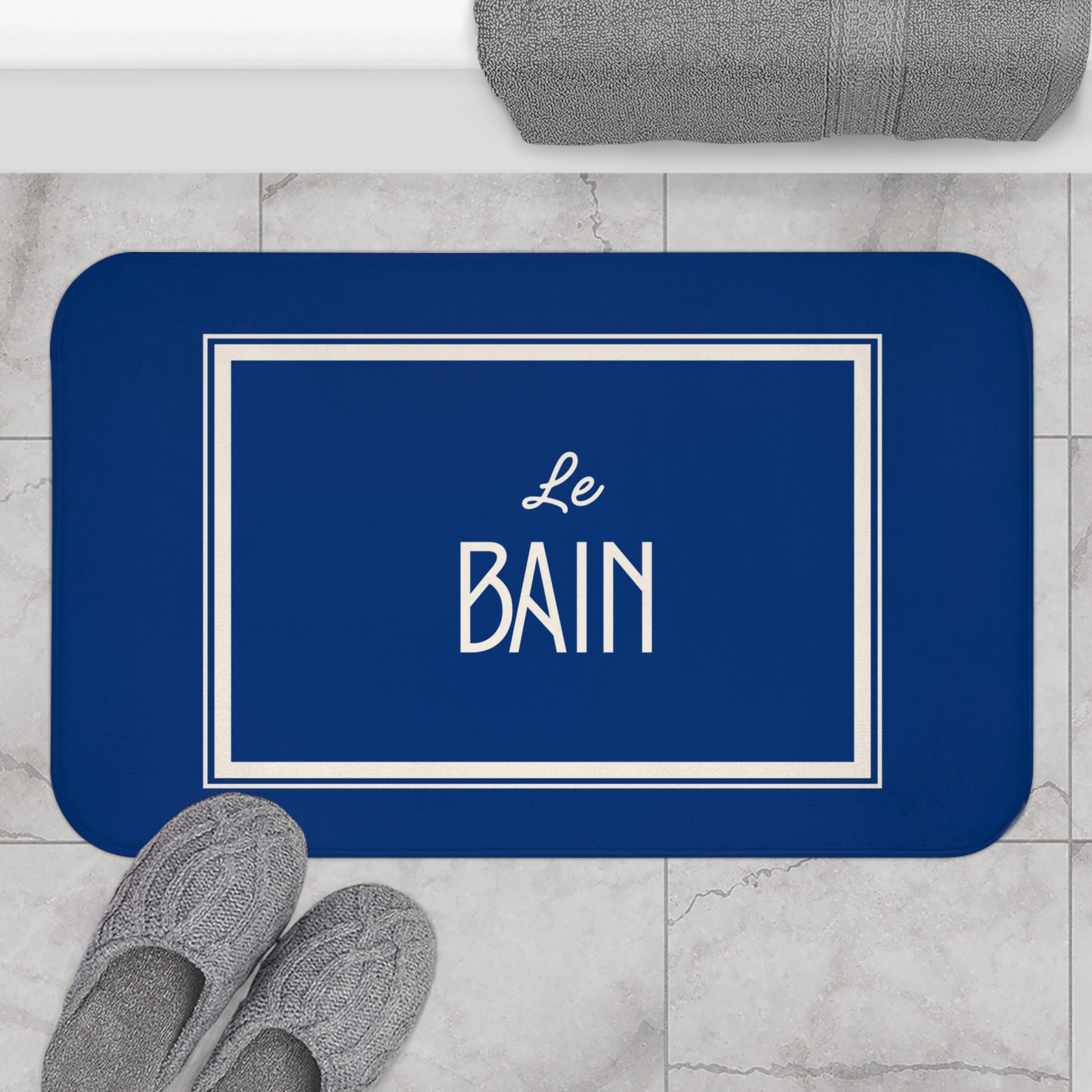 Blue bath mat with 'Le Bain' text on a marble floor with slippers and towel.