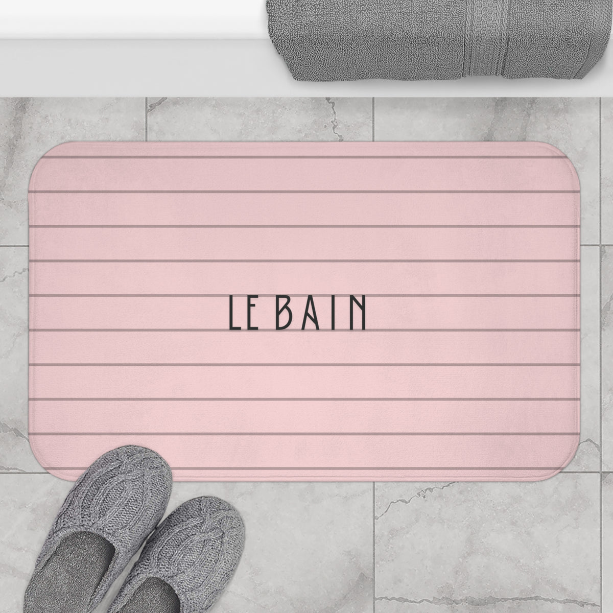 A pink bath mat with horizontal lines and the word 'LE BAIN' in the center, shown on a light marble tile floor with gray slippers and a gray towel nearby.