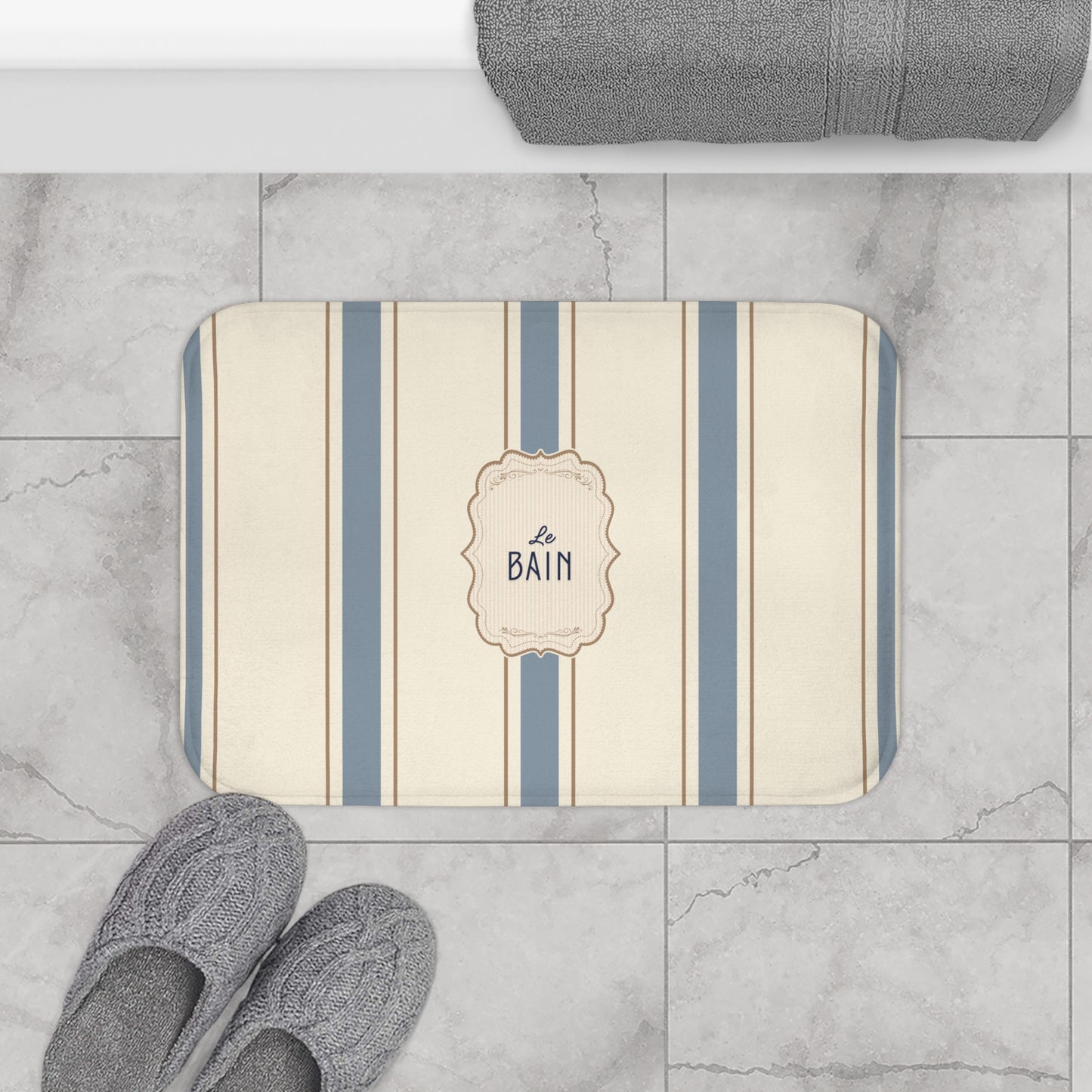 Striped bath mat with 'Le Bain' text on a marble floor with slippers and a towel.