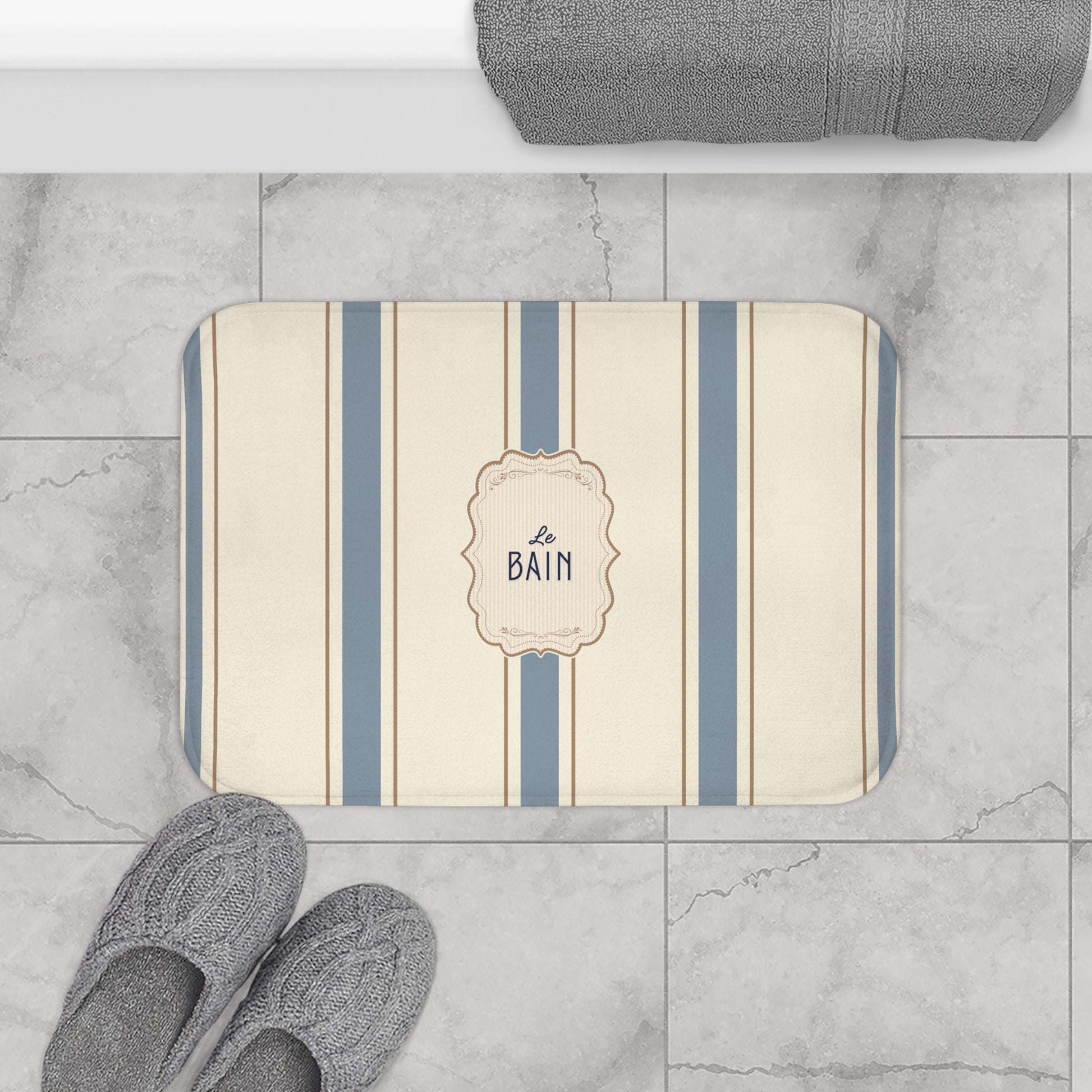 Striped bath mat with 'Le Bain' text on a marble floor with slippers and a towel.
