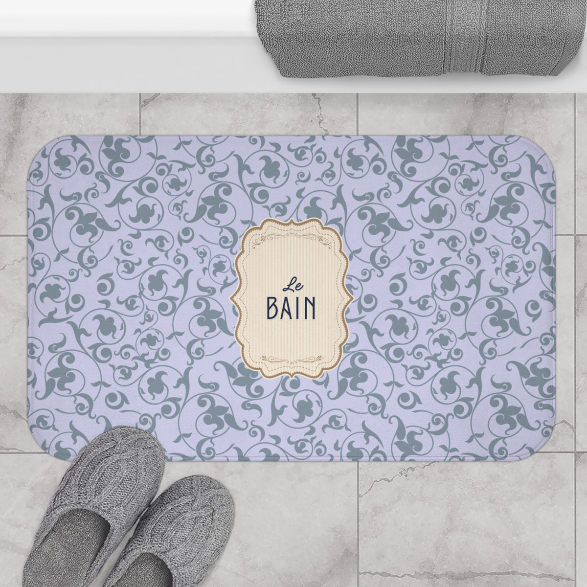 A rectangular bath mat with a lavender and gray damask pattern and a central cream label reading 'Le Bain', shown on a tiled bathroom floor with gray slippers and a folded towel nearby.