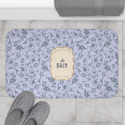 A rectangular bath mat with a lavender and gray damask pattern and a central cream label reading 'Le Bain', shown on a tiled bathroom floor with gray slippers and a folded towel nearby.