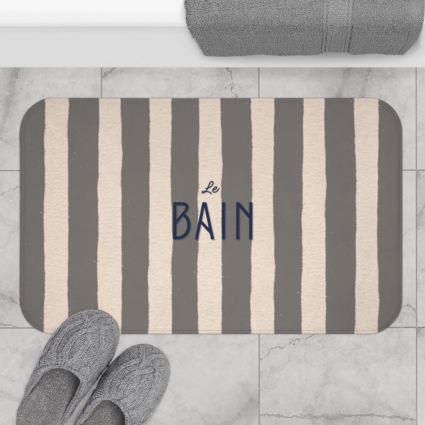 Striped bath mat with 'Le Bain' text on a marble floor with slippers and towel.