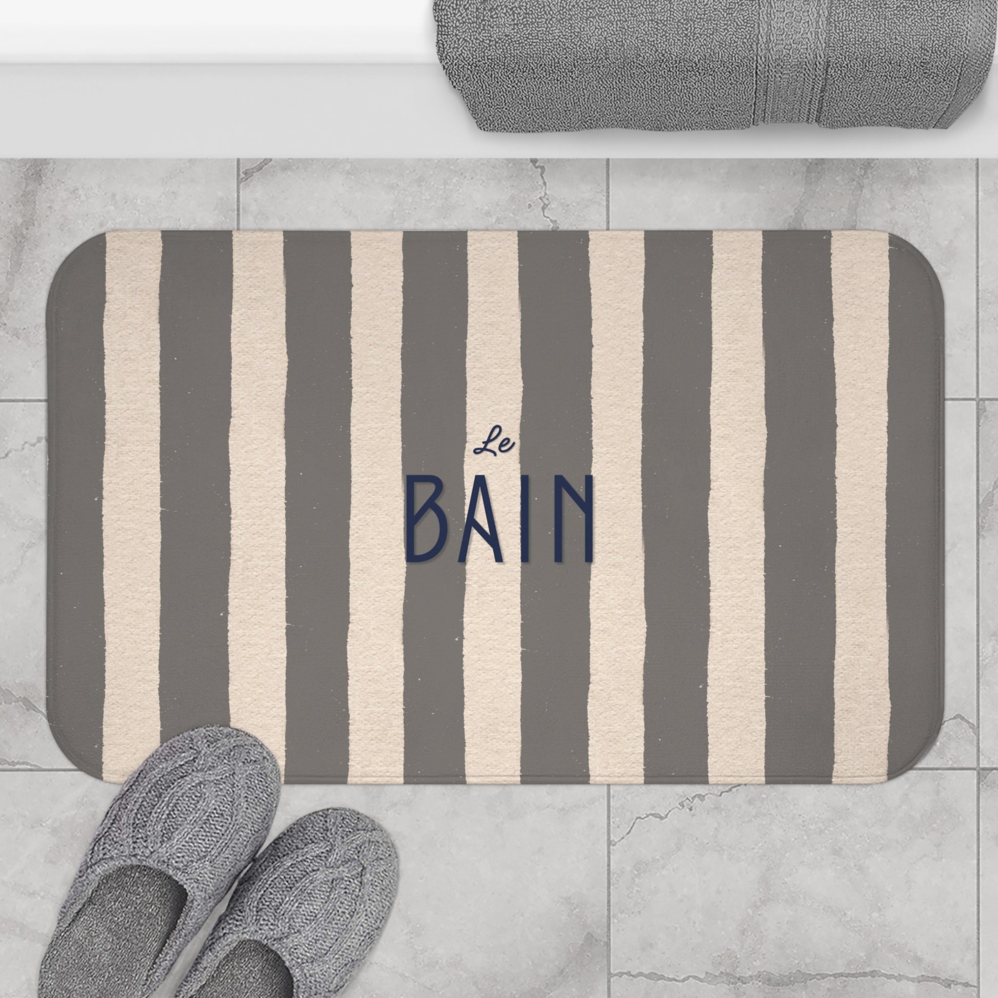 Striped bath mat with 'Le Bain' text on a marble floor with slippers and towel.