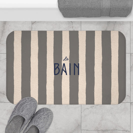 Striped bath mat with 'Le Bain' text on a marble floor with slippers and towel.