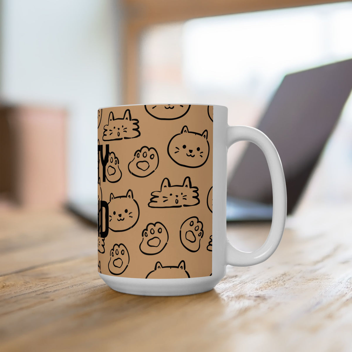 A white ceramic coffee mug with a tan background and black line-art cartoon cat faces, sitting on a wooden table with a blurred laptop in the background.