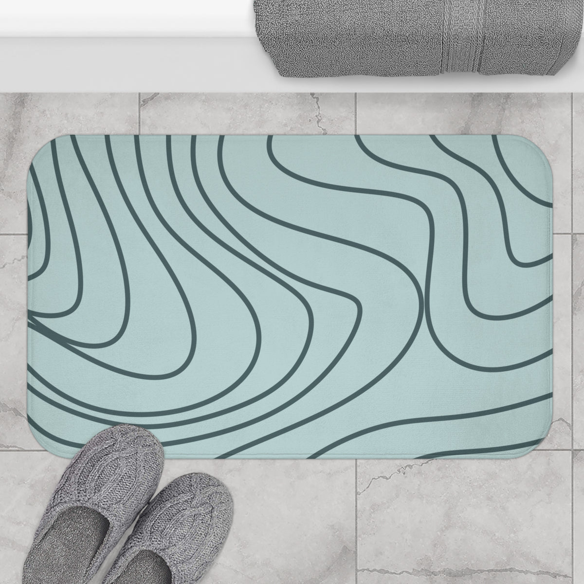 A rectangular bath mat with rounded corners featuring a light blue-green background and dark gray wavy contour-line pattern, shown on a tiled bathroom floor with gray slippers and a folded towel at the top.
