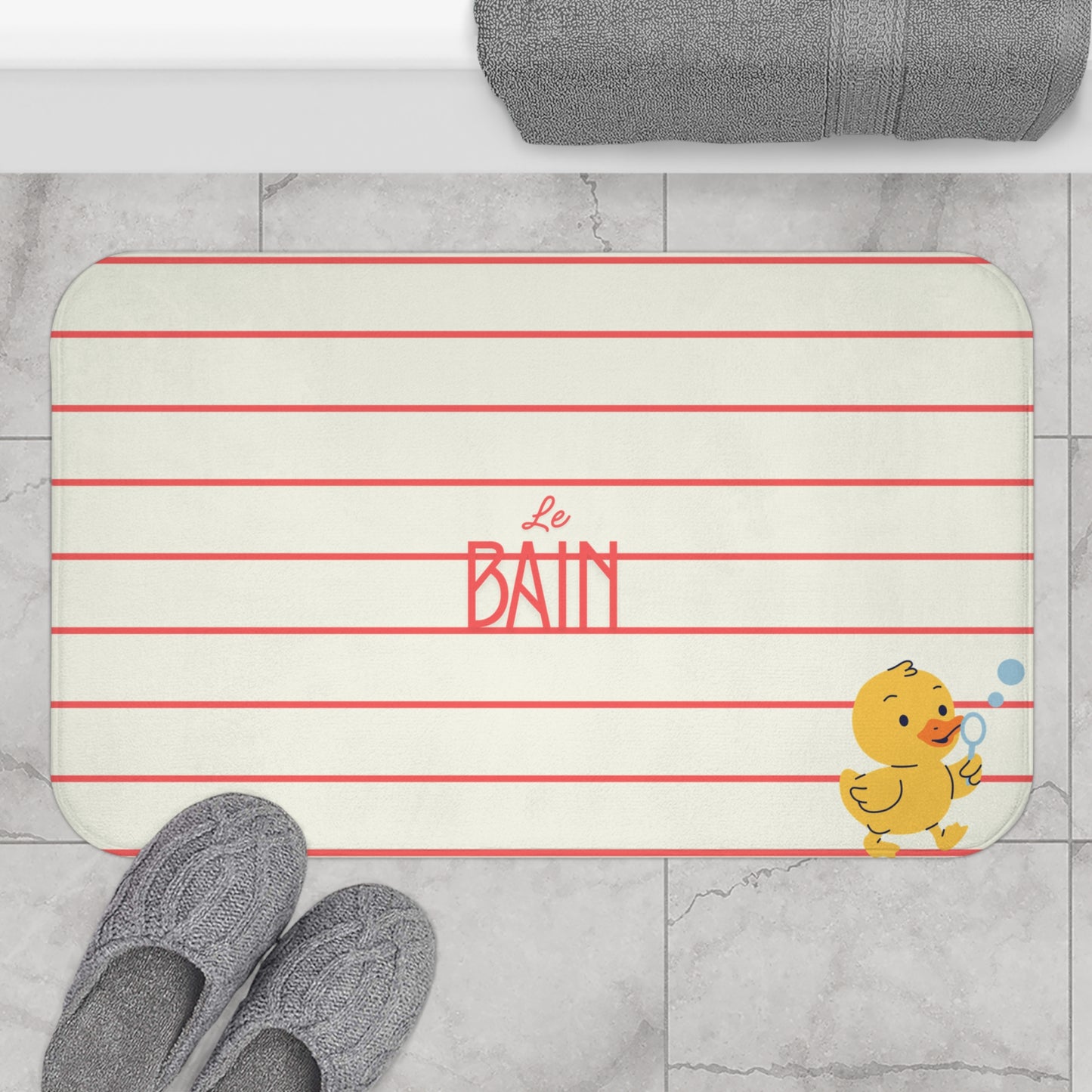 A rectangular bath mat with rounded corners, featuring horizontal red stripes on a warm off-white background, the words 'Le Bain' in red script and block lettering, and a small yellow duck holding a blue bubble wand in the bottom right corner. The mat is styled on a light gray marble floor with gray knit slippers and a folded gray towel visible.