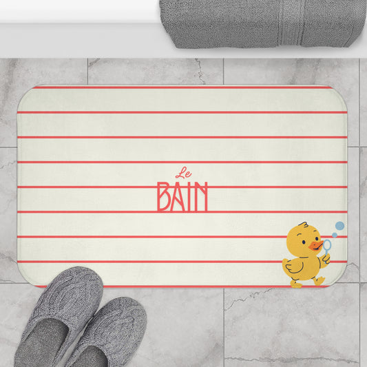 A rectangular bath mat with rounded corners, featuring horizontal red stripes on a warm off-white background, the words 'Le Bain' in red script and block lettering, and a small yellow duck holding a blue bubble wand in the bottom right corner. The mat is styled on a light gray marble floor with gray knit slippers and a folded gray towel visible.